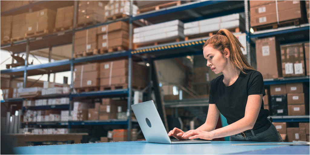 A woman standing in a warehouse and typing on a laptop placed on a large table.
