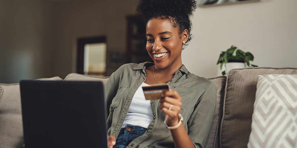 A woman of color sitting on a sofa with a laptop on her lap holding a payment card.
