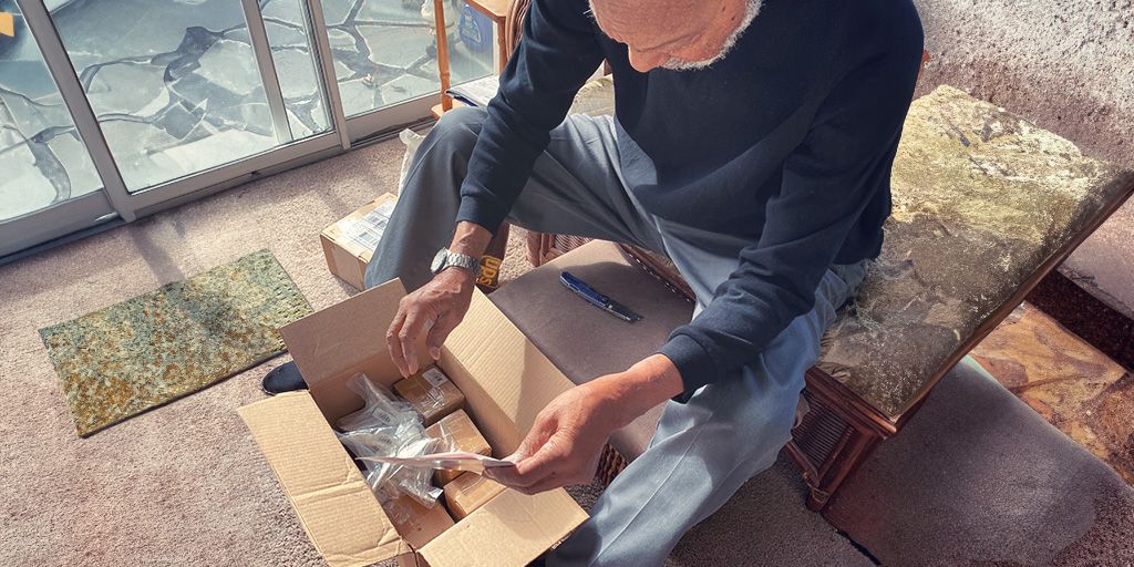 A man with a gray beard looking at box full of packages and holding a sheet of paper in his hand. 
