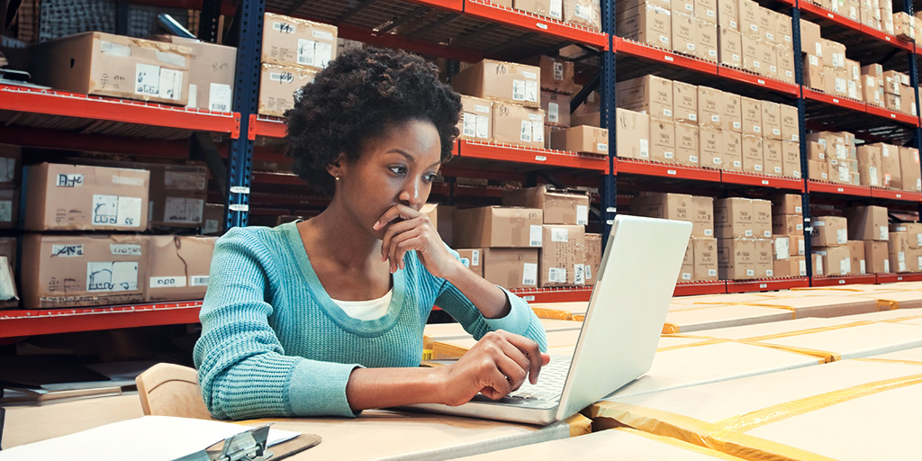 A woman of color sitting at a table in a warehouse. She is looking at a laptop that stands on the table.