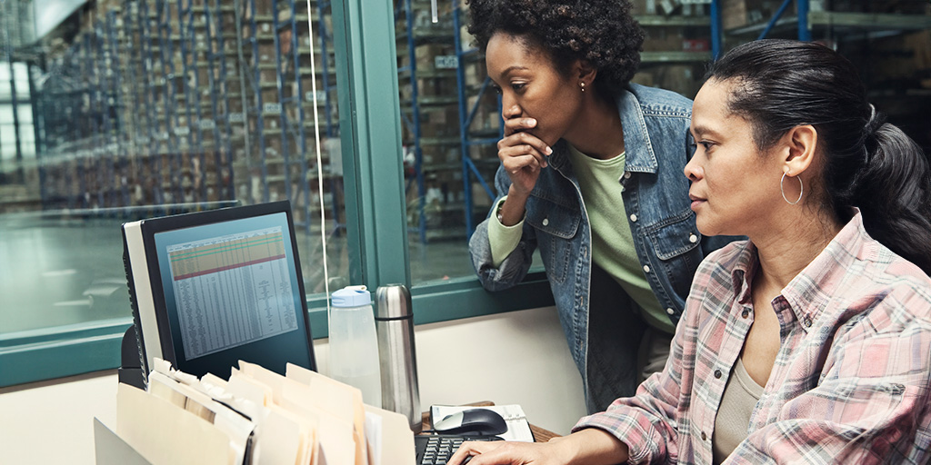 Two women of ethnic backgrounds look concerned at a computer monitor with a spreadsheet on it.