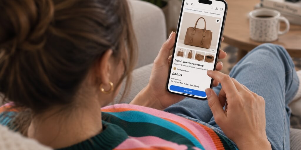 A woman sitting on a couch holding a smartphone in her hand. She is looking at an eBay listing of a handbag on the Smartphone.